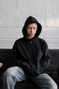 Teenager in a black hoodie sitting on a couch, reflecting on thoughts indoors.