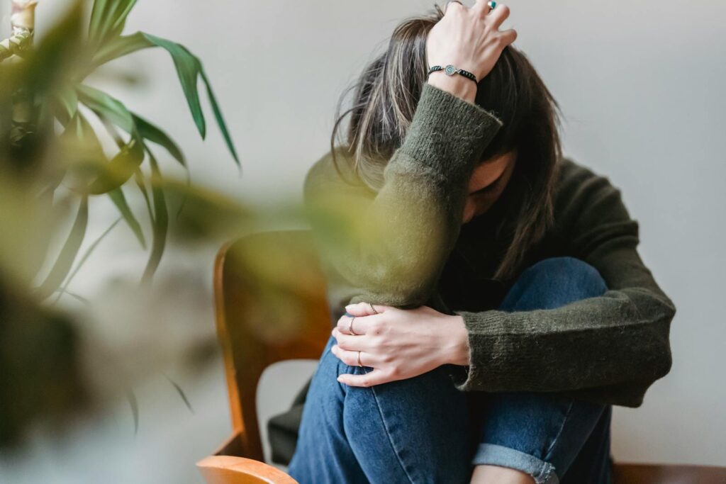 Anonymous young sorrowed female in casual outfit touching dark hair and embracing knees while sitting on chair at home